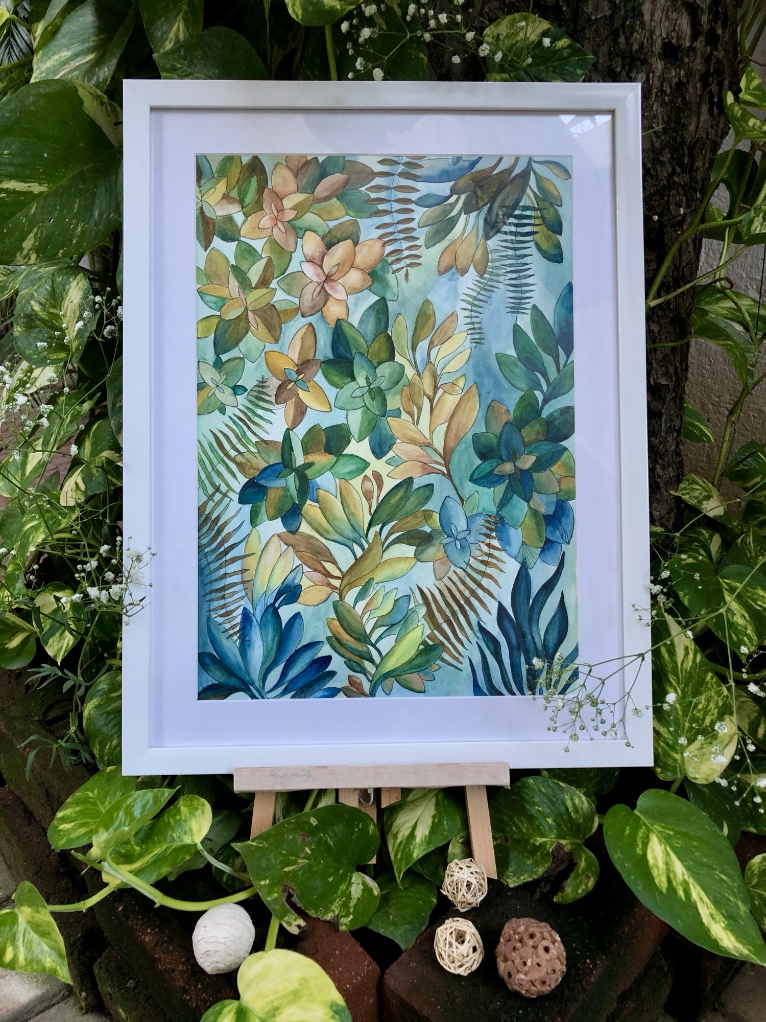 Framed watercolour painting of layered green and gold leaves and ferns, displayed among real potted plants