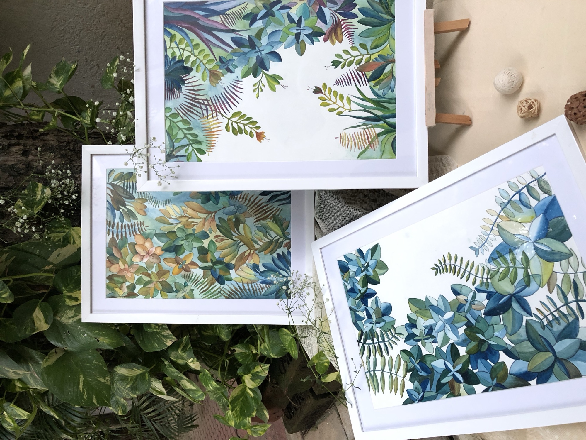 Three framed watercolour paintings of tropical leaves in blue, green and warm tones, laid out among potted plants